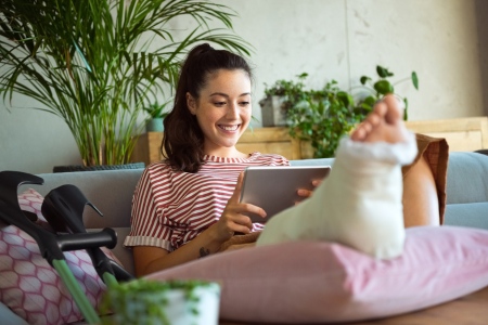 Eine Frau mit Gips am Bein sitzt auf der Couch und guckt lächelnd auf ein Tablet.